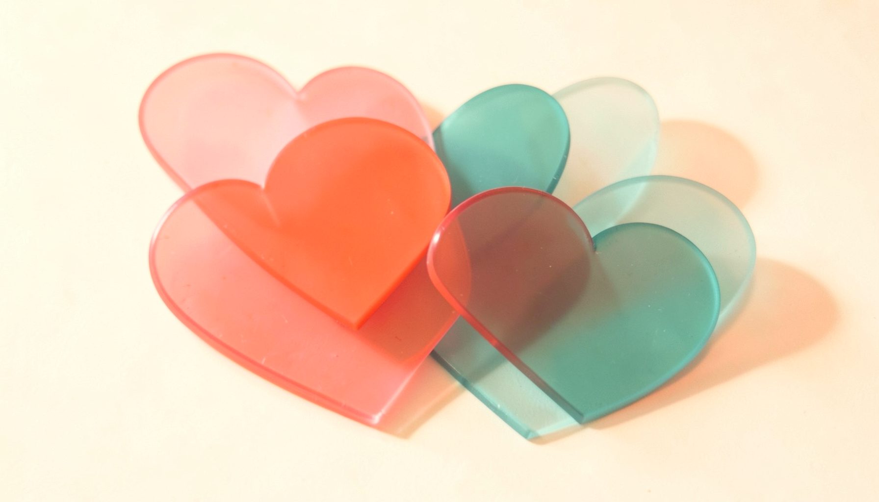 A warm, softly lit composition of overlapping translucent hearts in coral pink and teal against a creamy white background